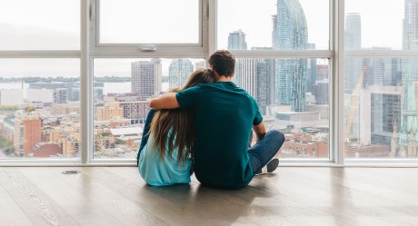 Ein Paar - Mann und Frau - von hinten, sie sitzen auf einem Holzfußboden vor einer großen Fensterfront. Im Hintergrund die Skyline einer großen Stadt mit vielen Hochhäusern.