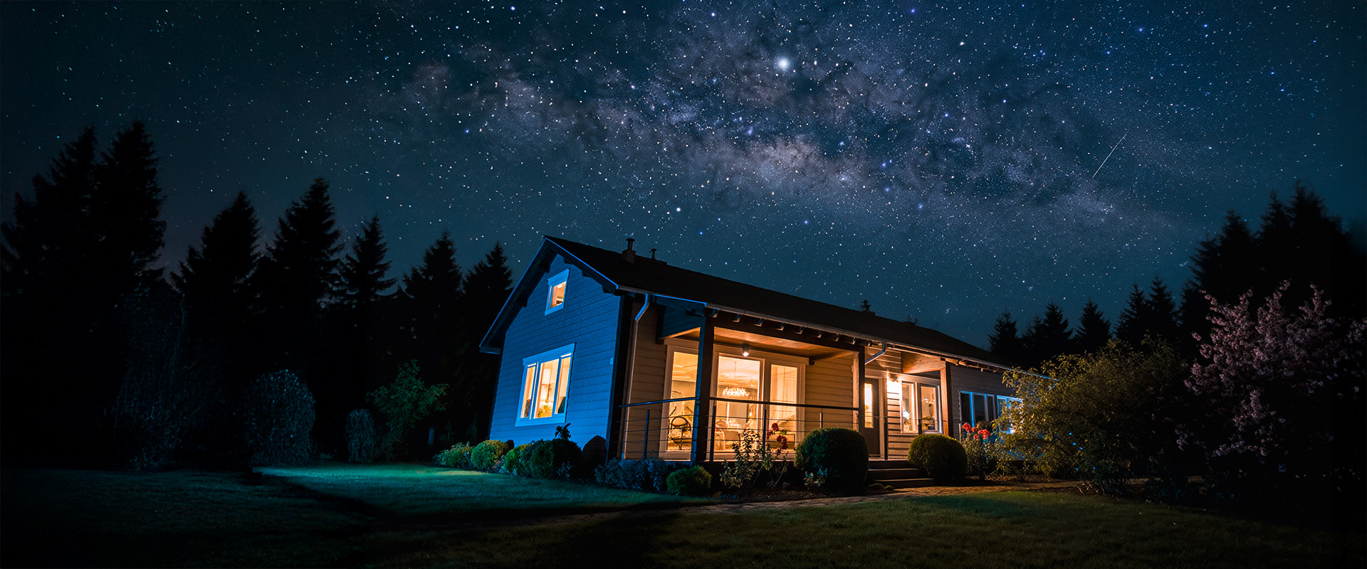 Eine Nachtaufnahme eines Einfamilienhauses in ländlicher Gegend über dem die Milchstraße am Himmel sichtbar ist.