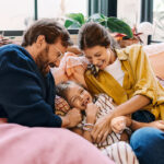 Vater und Mutter sitzen lachend mit Kind auf dem Sofa.