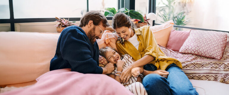 Vater und Mutter sitzen lachend mit Kind auf dem Sofa.