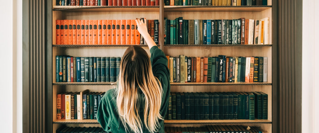 Eine Frau steht mit dem Rücken zur Kamera und holt ein Buch aus dem Bücherregal vor ihr.