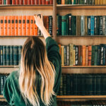 Eine Frau steht mit dem Rücken zur Kamera und holt ein Buch aus dem Bücherregal vor ihr.