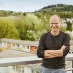 Mann steht am Geländer einer Dachterasse mit Wohnhäusern und Bäumen im Hintergrund.