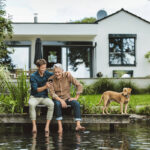 Mann mittleren Alters sitzt mit einem Mann im Seniorenalter vor einem Einfamilienhaus an einem Steg am Wasser, daneben ein Hund.